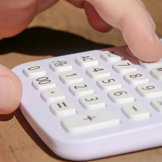 A hand pressing keys on a white calculator placed on a wooden desk in bright daylight.